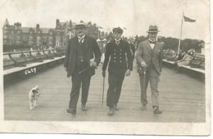 The young Reg looks dapper in his Merchant Navy uniform, carrying a baton and smoking a cigarette from the corner of his mouth, like his father William on the left. On the right, with a brolly, is his Uncle Alf. As his father is wearing a straw boater, they seem to be on holiday, striding along a wooden pier with women in white hats and a hotel in the background.
