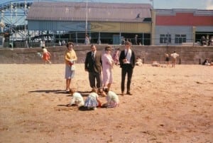 Reg and Thelma (centre), with Peggy and John Dale on the beach outside Wonderland, Cleethorpes, watch family members build sandcastles. Men wear ties. Note the wooden roller coaster, long demolished, where John had holiday work